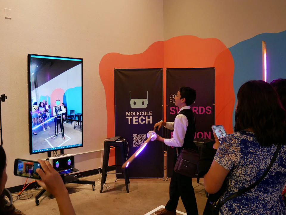 A visitor using an illuminated bokken beside the vertical body-tracking screen at PixelPerf Market.